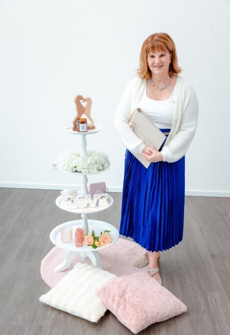 Traurednerin Susanne Schagen steht in einem eleganten blau-weißen Kleid am festlich geschmückten Trauplatz, bereit, eine emotionale Zeremonie zu leiten. Im Hintergrund sind stilvolle Dekorationen und eine romantische Kulisse zu sehen.